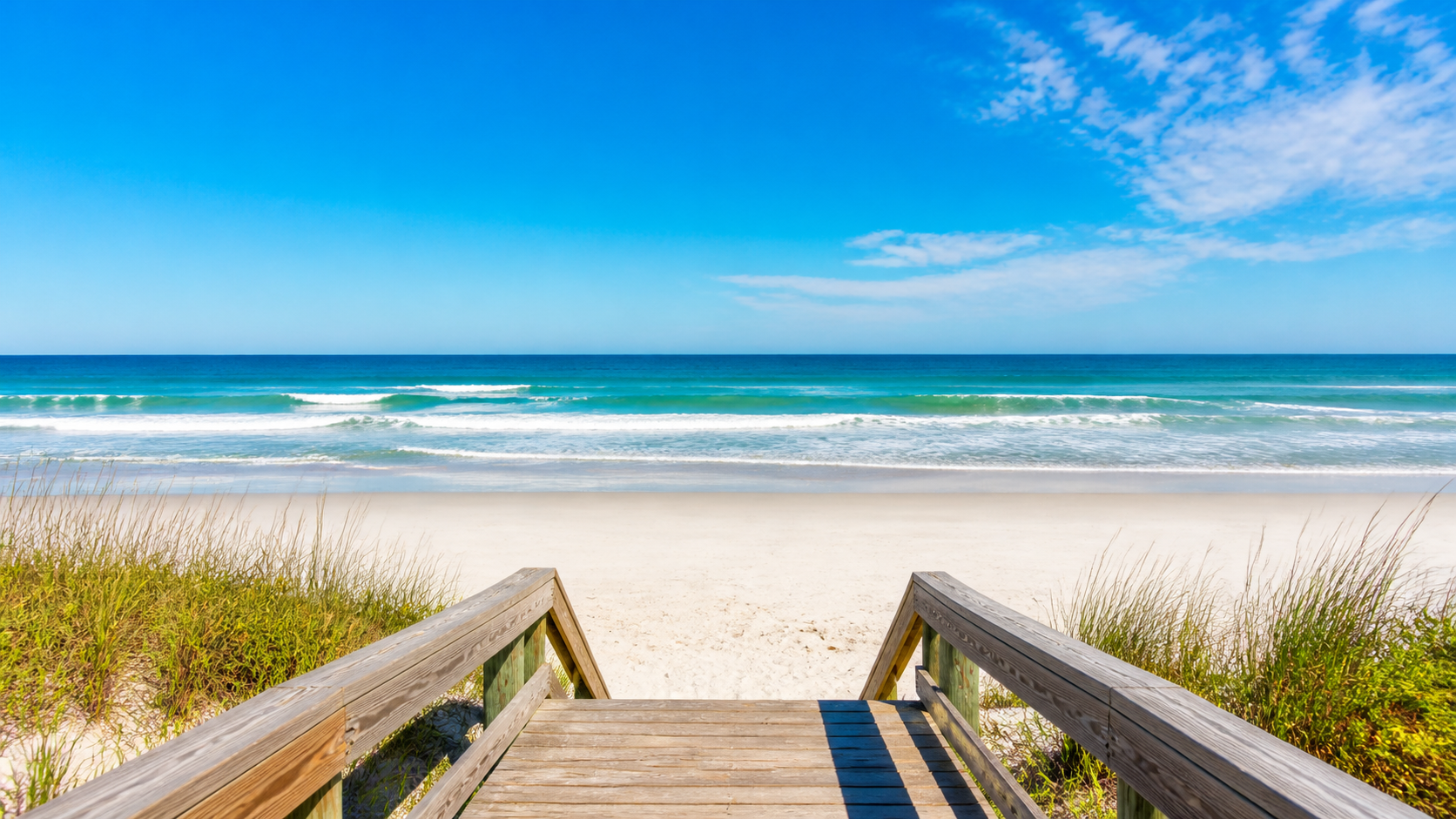 Boardwalk view facing white sand, ocean waves, and blue sky